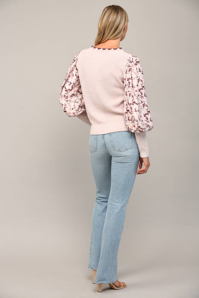 Woman wearing a light pink sweater with floral sleeves and light blue jeans on a gray background