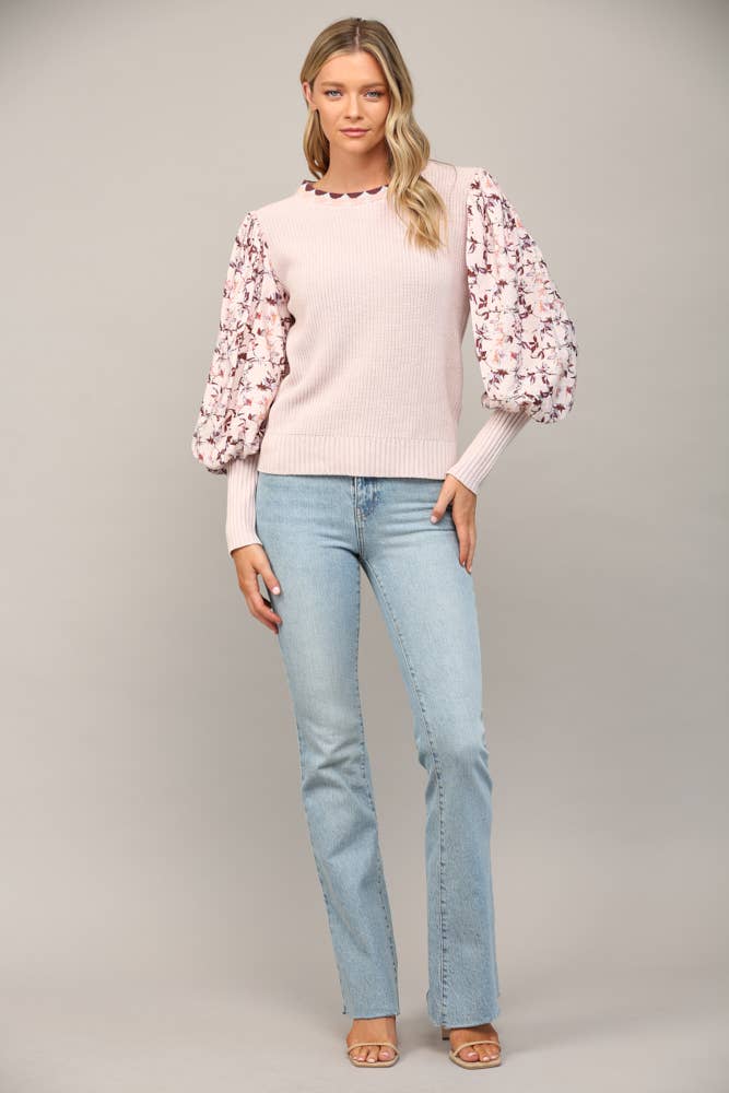Woman wearing a light pink sweater with floral sleeves and light blue jeans on a gray background