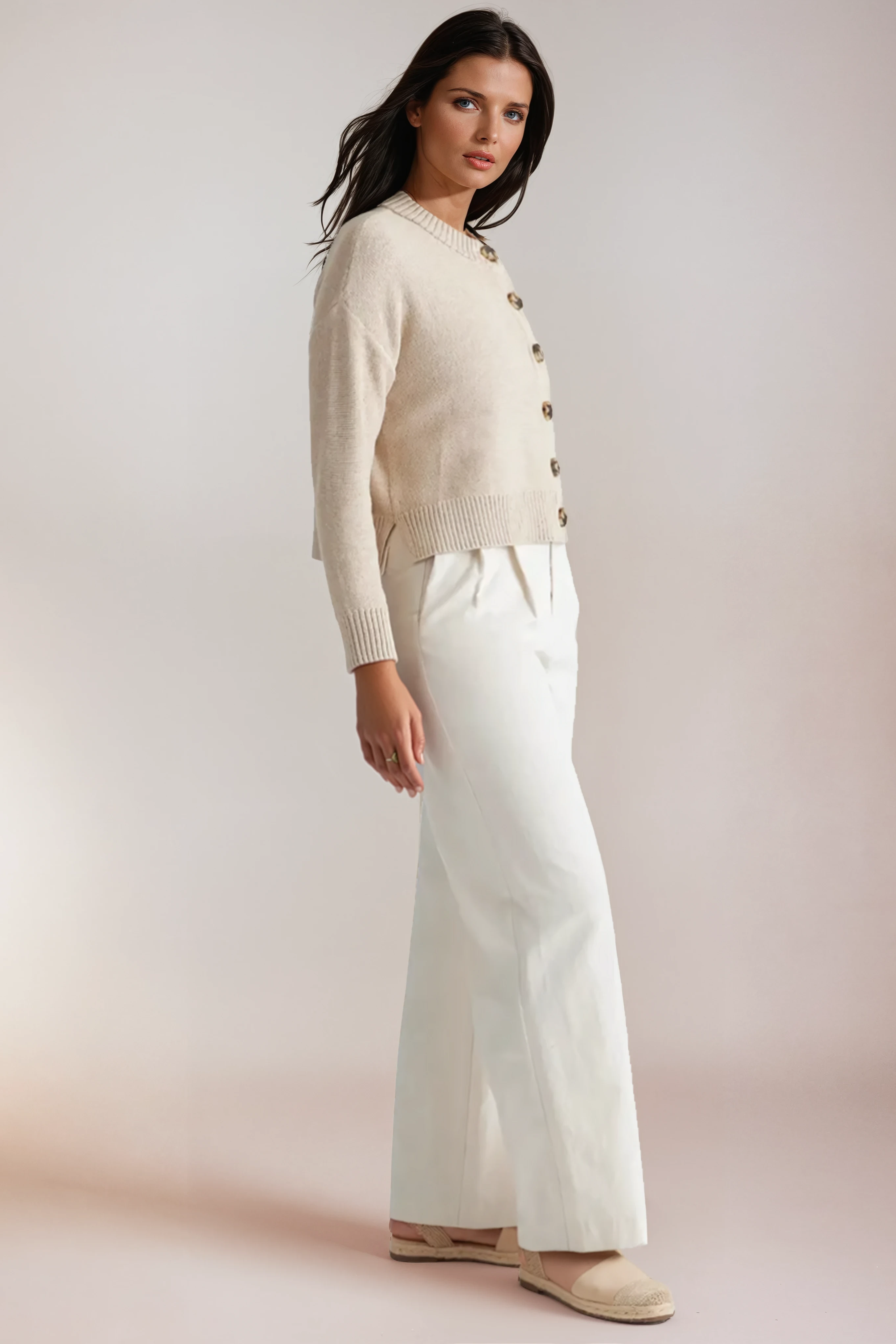 Woman wearing a beige cardigan and white pants against a plain background