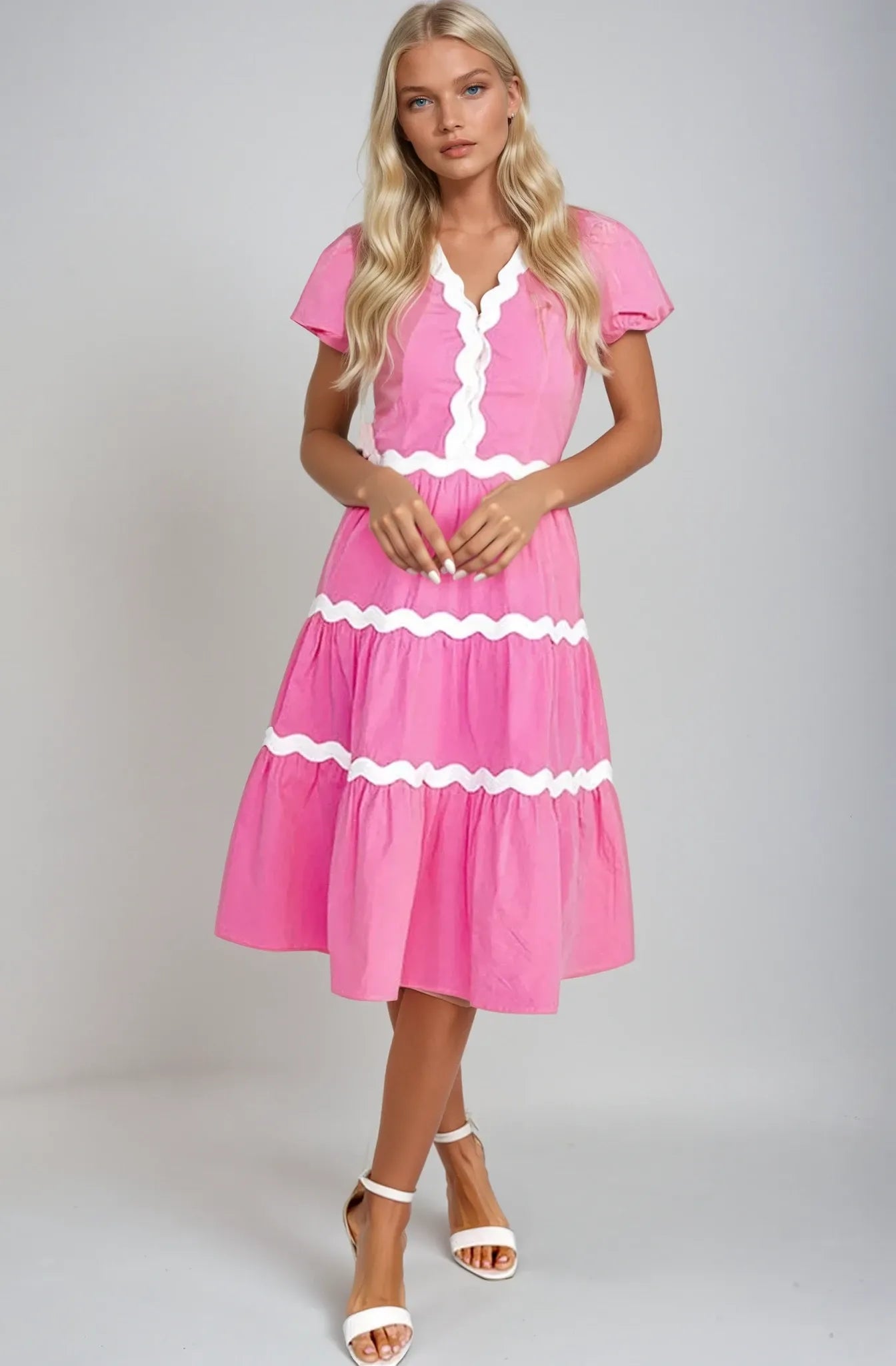 Blonde girl wearing a Cotton Pink Midi Dress with White Ric Rac Trim. Girl is in white studio wearing white high heel sandals. Girl holds her hands in front of her while staring into camera. Women's dresses, midi dress, cotton dress, summer dress, 100% cotton dress.