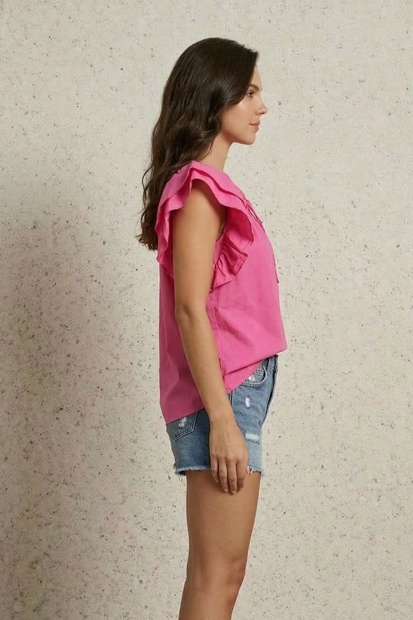 Woman wearing a pink top with ruffled sleeves and denim shorts against a neutral background